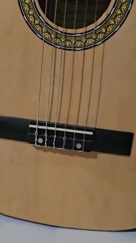 The camera focuses on the strings and body of a tan-colored classical acoustic guitar. The strings stretch from the black bridge, up past the sound hole, and toward the guitar's headstock. The sound hole is surrounded by a decorative ring of patterned concentric circles. The camera zooms in slightly on the sound hole, where a label is visible inside. The camera briefly shows the person holding the guitar and laughing at the end.