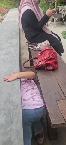 A young child wearing a pink floral t-shirt and blue jeans is shown partially concealed within a narrow gap between a wooden bench and a concrete wall. The child's head and torso are not visible, but their arms and legs are. One of the child's hands is extended upwards, resting on the concrete wall. On the wooden bench, a red handbag with a textured surface is placed near the child. Behind the bench, a woman wearing a black hijab and a black garment is seated, looking at a mobile device. The environment appears to be outdoors, with greenery and outdoor furniture visible in the background. The child appears to be exploring or playing within the gap.