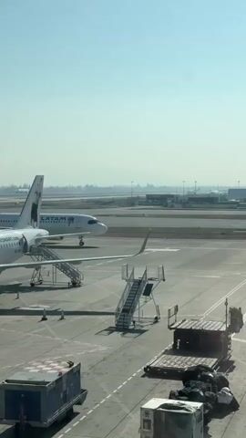 The video shows an airport tarmac on a clear, sunny day. In the foreground, there are airport service vehicles, including a baggage cart loaded with luggage and a blue container on wheels. To the left, a set of mobile stairs is positioned next to an airplane. In the mid-ground, a white airplane with the "LATAM" logo is visible on the tarmac. It appears to be taxiing or preparing for departure, with its engines possibly running. In the background, other airport infrastructure, including buildings and distant aircraft, can be seen under a bright sky. The overall scene depicts a typical operational environment at an airport.