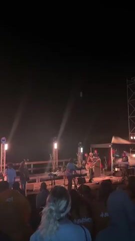 This is a vertical, low-angle, nighttime video of an outdoor concert. The stage is dimly lit with several vertical lights casting beams into the dark sky. A band with at least five members is performing. The person in the center foreground appears to be playing an acoustic guitar and singing into a microphone. To their left, another person is seated at a keyboard. To the right of the singer, other band members are playing instruments, including what appears to be a saxophone. The audience is seated or standing in front of the stage, with many people's backs visible to the camera. They are wearing casual clothing, and some have on hoodies. The overall atmosphere is that of a live music performance.