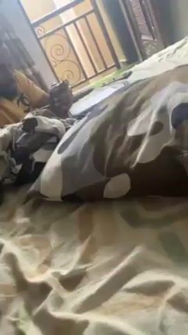 A man is lying in bed, looking at his phone, with a notebook or book next to him. The bed is covered in a patterned sheet and pillow. A metal railing is visible in the background.