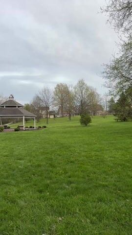 The video opens with a wide view of a manicured green lawn under a cloudy, overcast sky. A gazebo with a multi-tiered roof and white pillars sits at the edge of the lawn. Beyond the lawn, several houses are visible, partially obscured by trees with budding leaves. As the video progresses, the camera slowly pans to the left, revealing a sidewalk and a line of trees and bushes along the edge of the lawn. A small figure on a bicycle can be seen briefly in the middle of the lawn. The sound of a young child can be heard saying, "Da," and making other vocalizations.
