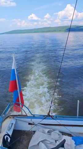 A video shows the wake of a boat moving through the water. The boat has a Russian flag attached to its railing. The water is blue and calm, with green hills in the distance under a partly cloudy sky. The boat appears to be moving forward, leaving a foamy trail behind it.