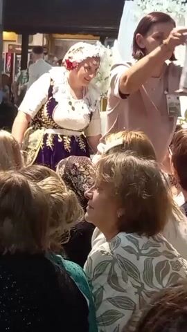 A woman in traditional dress, adorned with a headdress and jewelry, is interacting with a group of people. She holds a small, transparent pouch filled with what appears to be money or small items. Another woman, to her right, is also holding a similar pouch. The background suggests an indoor setting, possibly a market or a festive gathering, with glimpses of other individuals and decorative elements. The overall atmosphere seems to be one of a cultural celebration or exchange.