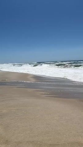 The video depicts a serene beach scene at Torreira, Portugal. The camera is positioned on the sandy shore, looking out towards the vast ocean. The sky is a clear, vibrant blue, indicating a bright and sunny day. Gentle waves roll in from the sea, creating a frothy white surf that washes onto the beach. The water appears clear and inviting, with the sunlight reflecting off its surface. The sand is a warm, light brown, with subtle textures and patterns visible from the receding waves. There are no people or man-made objects in the frame, emphasizing the natural beauty of the location. The overall mood is peaceful and tranquil, evoking a sense of calm and relaxation. The video captures the timeless rhythm of the ocean, with the continuous ebb and flow of the tide. The time of day appears to be midday, given the bright lighting and clear sky. The location is explicitly stated as Torreira, Portugal, a coastal town known for its beaches. There is no visible text within the video itself.