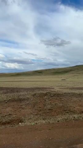 This media file captures a scenic view of a vast, rolling landscape under a partly cloudy sky. The foreground is dominated by a dry, reddish-brown dirt road or path, with scattered pebbles and sparse, dry grass. Beyond the road, a wide expanse of dry, golden-brown grass stretches towards a gentle, undulating hillside. This hillside is covered in similar dry vegetation, with patches of darker green or brown suggesting variations in the terrain or plant life. Towards the top of the hill, there are visible rock formations or a cluster of larger rocks, adding some texture to the distant horizon.

The sky above is a dynamic mix of bright blue patches and significant cloud cover. Large, fluffy white clouds are interspersed with darker, heavier clouds, creating a dramatic and somewhat moody atmosphere. The lighting suggests it is daytime, possibly late morning or early afternoon, given the brightness of the illuminated clouds and the contrast with the shadowed areas.

There are no people or man-made objects prominently featured in the scene, aside from the dirt path. The overall impression is one of a remote, natural environment, possibly a steppe or prairie. The activity taking place is implied to be a journey or travel along the dirt path, with the camera moving slightly, causing a subtle shift in perspective. The location cues from the prompt, "Bakty, Kazakhstan," suggest a dry, open, and potentially arid environment, which aligns with the visual evidence. The mood is one of expansive natural beauty, with a touch of wildness and solitude. No visible text is present in the media.