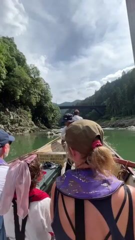A group of people are on a boat traveling down a green river. The boat has a person steering it in the front. The people in the boat are wearing casual clothing and life vests. The river is surrounded by lush green trees and rocky cliffs. There is a bridge in the background. The sky is cloudy with patches of blue. The camera is mounted on the boat and facing forward.