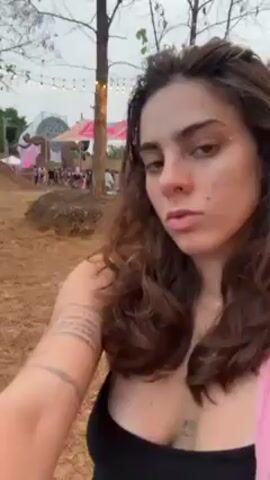 A woman is standing outdoors.  She is wearing a black tank top and a pink shawl.  She has brown hair and a tattoo on her arm.  The camera is held up close to her face.  In the background there are other people, lights, and structures.  She then lies back in a pink hammock strung between wooden poles.  The camera pivots, revealing a lush green landscape beyond the edge of the cliff where the hammock is strung.  Another person is briefly visible at the edge of the frame, wearing a blue top.