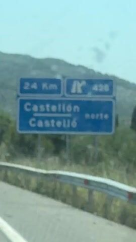 The video shows the view from inside a vehicle traveling on a highway. A blue road sign is visible in the distance, indicating directions to Castellón and Castelló. The sign also shows distances and highway numbers. Sunlight is streaming through the windshield, and the sky is clear. The interior of the car is visible in the foreground, including the dashboard and the legs of a passenger. The scenery outside consists of rolling hills and trees.