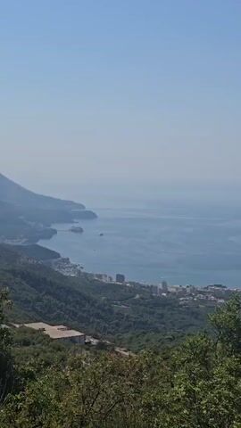 The video shows a panoramic view from a mountaintop overlooking the sea. In the foreground, there are trees and lush green vegetation. The midground reveals a coastal town with buildings nestled on the hillsides, and a body of water with a few boats. Further out, the vast expanse of the sea stretches to the horizon under a clear blue sky.
