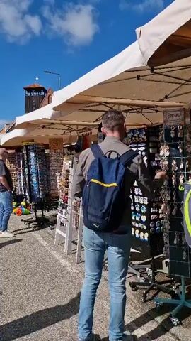 A man wearing a blue backpack and jeans walks past stalls selling souvenirs. The stalls are covered by white canopies. The man stops to look at a display of magnets and postcards. The sun is shining and the sky is blue with some clouds.