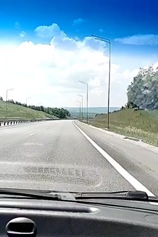 This is a first-person perspective video taken from inside a vehicle moving along a highway. The road has multiple lanes and is bordered by grassy embankments and trees. Streetlights are visible along the right side of the road. The sky is bright blue with fluffy white clouds, indicating fair weather. The dashboard of the vehicle is partially visible in the foreground. The video appears to be a short clip from a longer journey.