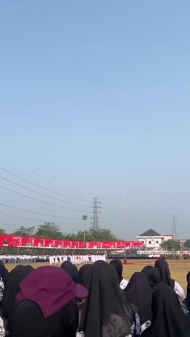 This video shows a crowd of people, mostly women wearing black hijabs, viewed from behind, facing a wide open field under a clear blue sky. In the distance, a line of people in uniform stands at attention. A red banner with white lettering is stretched across the field, and power lines and towers are visible in the background. The overall scene suggests an outdoor ceremony or event.