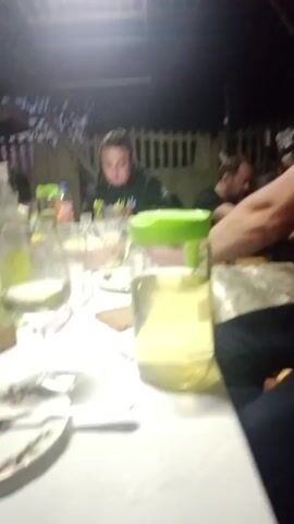 The video shows a group of people gathered around a table outdoors at night. In the foreground, a man in a black t-shirt and jeans is sitting and looking down at something on the table. To his left, a woman in a green hoodie is also seated and looking at the table. Further back, another man is visible. A pitcher of yellow liquid, plates, glasses, and food are on the table. The background shows a fence and some foliage. The lighting is dim, suggesting it is evening or night.