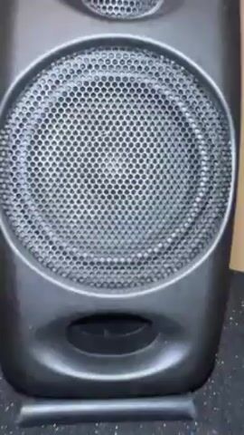 The video features a close-up of a dark grey speaker. The speaker has a large, circular grill covering the main driver, with a smaller tweeter above. Below the main speaker is a bass port. The speaker sits on a speckled black surface. The music is playing, causing the speaker to vibrate slightly.