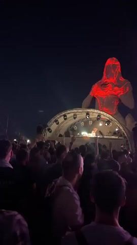The video shows a nighttime outdoor concert or festival. A large crowd of people are gathered in front of a stage. On the stage, a DJ is performing with turntables and a laptop. Behind the stage, a massive inflatable sculpture of a figure with glowing red internal lights is illuminated against the dark sky. A small airplane is visible flying in the distance above the sculpture. The overall atmosphere appears energetic and festive, with the crowd raising their hands in the air.