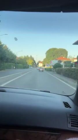 The video shows a view from inside a car driving on a road. The camera is pointed forward through the windshield. The road is paved and appears to have at least two lanes in each direction. There are other cars on the road, traveling in the same direction as the camera car. On either side of the road, there are trees, hedges, and buildings, including what appear to be houses and businesses. The sky is clear and blue, and the lighting suggests it is daytime, possibly late afternoon or early evening. The dashboard of the car is visible in the foreground.