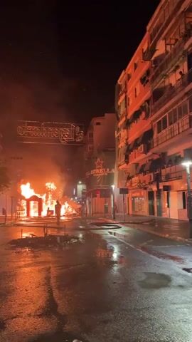 The video depicts a street scene at night, illuminated by a large fire in the center. The fire appears to be consuming a structure, possibly a temporary installation or float, given the presence of an arched entryway and what looks like decorative elements. Smoke billows into the night sky. The street is wet, with reflections of the fire on the asphalt, suggesting it may have rained or been recently cleaned. Several figures, appearing to be people, are visible in the background, silhouetted against the flames, some seemingly observing the event. Buildings line both sides of the street, some with lit windows or signs, adding to the urban nighttime atmosphere. A banner with text and a star logo hangs from one of the buildings to the right of the fire. The overall impression is one of a dramatic event, possibly a festival or a controlled burn, taking place in a public urban space.