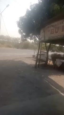 A roadside scene with a small food stall sign visible. The sign reads "WARUNG SATE BLENDO" and "BU ISAH" with an address beneath. The stall has a roof supported by wooden poles. In the foreground, a dirt or gravel road is visible with dust or dirt kicked up, possibly from a vehicle. Trees are present in the background, and a truck is visible driving away in the distance. The lighting suggests it is daytime, possibly morning or late afternoon due to the low angle of the sun. There are no people clearly visible in the frame.