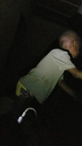 The video opens in a very dark environment, with a young child, likely a boy, lying down on their side. The child is wearing a light-colored (possibly white or light grey) short-sleeved t-shirt and dark bottoms. A bright white cable, resembling a charging cord with an illuminated end, is visible near their lower body, acting as a primary light source in the dimness. An adult voice softly says "Okay."

At 0:01, the child sits up, turning their head towards the camera with a smile. They raise both arms above their head in a playful or stretching gesture. The lighting briefly brightens, showing more of the child's face and upper body, and their short, dark hair.

The child continues to move their arms playfully and look towards the camera at 0:02, maintaining their cheerful expression. Another indistinct voice can be heard.

By 0:03, the child begins to lower their arms and moves as if lying back down or shifting out of the direct light. The scene darkens significantly, making the child's features less clear.

From 0:04 to the end, the video is almost entirely dark, with only faint outlines of the surroundings and a distant, small light source barely visible. The child is no longer distinctly in view.

The video captures a brief, playful moment with the child in a very low-light setting, likely illuminated by a mobile phone's flashlight, causing the fluctuating brightness.