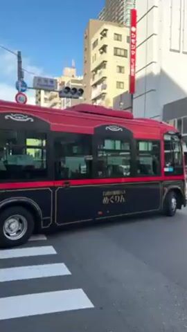 The video shows a bus, painted red and black, stopped at a crosswalk in a Japanese city. The bus has the text “めぐりん” written on its side. The bus’s license plate reads “37-05”. The crosswalk is at an intersection, and the traffic light is red. Various buildings and a tall red billboard with white text are visible in the background. A person on a bicycle rides through the crosswalk in front of the bus.