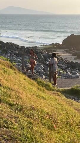 A young woman in a pink bikini walks down a wooden staircase, laughing. A young man in a gray shirt and khaki shorts stands facing her, looking at her and holding something in his hands. Behind them, a rocky beach leads to the ocean, where waves crash and several people are visible in the water. The sun is low in the sky, casting a warm glow over the scene. The camera pans across the landscape, capturing the beach, the ocean, and the surrounding vegetation.