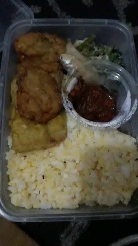 A close-up, top-down shot shows a clear plastic takeaway container filled with food. The container is divided into sections. The largest section is filled with white rice. To the left of the rice, there are several pieces of fried food, appearing to be tempeh. Above the rice and to the right, there is a small, clear plastic container with a silver foil lid, holding a dark red chili paste. Next to the chili paste, there is a small fish and some green vegetables. The background appears to be a dark, patterned surface, possibly fabric. The lighting is dim.
