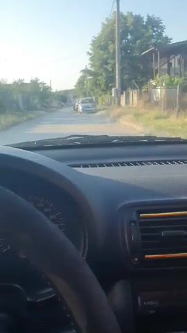 This is a first-person perspective video taken from inside a car, likely a Seat, as indicated by the logo on the steering wheel. The driver's hands are on the steering wheel, and the dashboard instruments are visible. The speedometer shows a reading of approximately 20-30 km/h, suggesting the car is moving at a moderate speed.

The scene outside the car is a rural or semi-rural road in Vaglen, Bulgaria. On either side of the road, there are houses, fences, trees, and vegetation. The sun appears to be shining, indicating it is daytime, likely late afternoon or early evening due to the soft, warm light. The weather seems clear and pleasant.

Several cars are visible on the road ahead, suggesting other people are also traveling in the area. The overall atmosphere appears calm and ordinary, depicting a typical drive through a village. The video does not contain any explicit text, but the dashboard instruments provide information about the car's status. The red warning light on the dashboard might indicate a minor issue with the car, but it doesn't seem to be causing immediate concern to the driver.