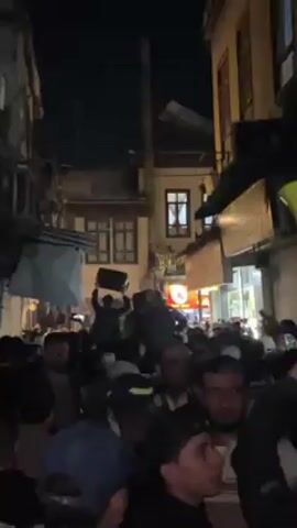 A crowd of people, likely men, is gathered in a narrow street at night. A few individuals are holding up a black object, possibly a loudspeaker, above their heads and dancing. The crowd appears celebratory, possibly due to music or an event. The street is lined with buildings; one is a light-colored, two-story structure with dark-framed windows and some decorative lighting. A sign with what appears to be a red circle is visible in the background. The video is shaky, taken from within the crowd.