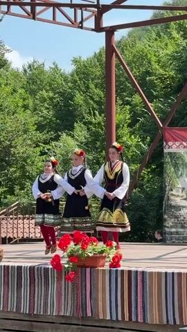 Three young women in traditional folk costumes are performing a dance on an outdoor stage. The costumes consist of black dresses with colorful embroidered trim, white blouses, and red and white striped socks. They are adorned with red flowers in their hair. The stage is decorated with a striped fabric runner and pots of red flowers. Behind the stage, lush green trees form a backdrop, and a metal structure supports a banner with Cyrillic text. The women are moving in unison, their arms extended and hands clasped. The lighting suggests a bright, sunny day.