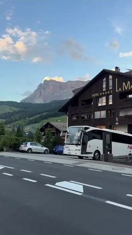 The video shows a street scene with a hotel and mountains in the background. In the foreground, there is a paved road with white lane markings. To the right, a large white tour bus is parked in front of a dark brown, multi-story hotel building with a sign that reads "HOTEL ***** LA MA". The hotel has several windows, some of which are lit. To the left of the hotel, there are two cars parked on the side of the road: a silver car and a smaller blue car. Beyond the cars, there are trees and more buildings, with a steep, forested mountainside rising behind them. In the distance, a large, rocky mountain peak is visible against a cloudy sky with patches of blue. The overall lighting suggests it is late afternoon or early evening.