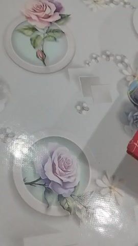 The video displays a close-up, overhead view of a light-colored surface decorated with a floral pattern. The pattern features a prominent circular frame containing a light purple rose with green leaves. Above this, another circular frame encloses a pink rosebud. Scattered across the surface are small, lighter-colored embellishments resembling blossoms or beads, as well as some faint square shapes. The surface appears to have a textured, possibly plastic or laminated finish, with noticeable glare from lighting. The camera angle is slightly tilted, and the video is a still frame.