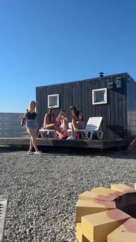 The video shows four young women in front of a small, dark cabin on a sunny day. The ground is covered in gray gravel. Three of the women are sitting on white lounge chairs on the cabin's small wooden porch. They appear to be drinking. The fourth woman is standing on the gravel in front of the cabin, holding a bag of chips. She gestures with one hand as she walks toward the cabin. The cabin is black with two small, square windows and a birdhouse on the side. A gray wooden fence is behind the cabin.