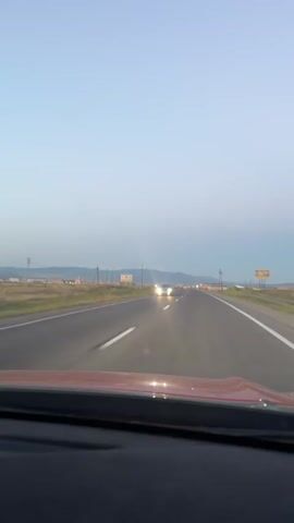 The video is from the perspective of a driver looking forward on a highway. The sky is a pale blue, suggesting either early morning or late evening. The highway has two lanes in each direction, separated by a white line. The road surface is asphalt and appears to be in good condition. To the left and right of the highway, there are flat, open fields with some scattered vegetation and distant buildings. There are also utility poles along the side of the road.

In the distance, on the opposing lanes of traffic, a car with its headlights on is visible. It appears to be a lighter-colored sedan. As the video progresses, other distant lights, possibly from other vehicles or buildings, can be seen along the highway. The video captures the car moving forward on the highway, with no other significant interactions or events occurring. The overall environment is rural and open, with the highway being the primary focus.