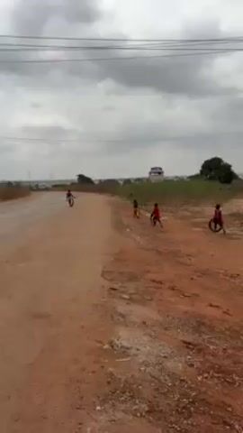 The video shows four children, possibly boys, playing on a dirt road in an open area. All the children appear to be holding or manipulating black tires or tire rims. The child farthest from the camera is running down the road away from the camera while holding his tire. The other three children appear to be standing or walking with their tires along the side of the road. The area is rural or suburban, with visible buildings in the distance and utility poles along the side of the road. The sky is overcast with grey clouds.