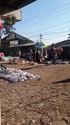 The video shows an outdoor market scene. In the foreground, there are piles of what appear to be goods covered in plastic sheeting on the dirt ground. In the middle ground, several people are gathered around stalls or tables, some with umbrellas for shade. One man in a blue shirt is walking away from the camera, carrying a bundle of items. The background consists of buildings that appear to be shops or market stalls, with power lines visible overhead against a clear blue sky. The overall environment is dusty and appears to be a typical open-air market.