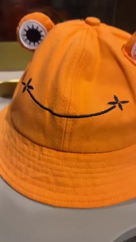 The video shows an orange bucket hat with frog eyes and a smiling face placed on a table. Behind the hat is a glass enclosure with a large, reddish-brown, textured object inside, possibly a replica of a cacao pod. The enclosure is part of a larger display that resembles a tree trunk with an opening. In the background, there is a sign with instructions that include a hand symbol and a prohibition against touching or sitting on a fountain. Further in the background, a blurry image of a person can be seen. The environment appears to be indoors, likely a museum or exhibition.
