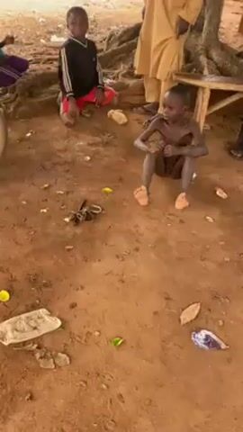 The video shows a scene set outdoors on a brown, dirt-covered ground. Three children are present, two seated and one partially visible to the left of the frame. One child is kneeling, wearing a long-sleeved black shirt and red shorts, pointing off-camera to the right. Another child is sitting, shirtless and covered in what appears to be dirt or mud. An adult, partially visible to the right, is wearing a beige or light brown robe. To the left, part of a motorcycle is visible. There is trash, scattered debris, and leaves on the ground. In the center of the frame is a chain and a few keys. The overall lighting is muted, suggesting an overcast day or a shaded area.