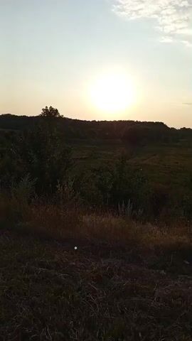 The video shows a landscape at sunset. The sun is low in the sky, casting a warm glow over the scene. There are trees and bushes in the foreground and background. In the middle of the video, two people are seen riding bicycles on a road. One person is wearing a red shirt. The camera is moving, giving a sense of motion. The scene appears to be rural or semi-rural. The lighting suggests it is either dawn or dusk.