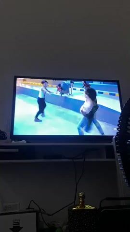 This video captures a scene from a video game displayed on a television screen. The game appears to be a fighting game or a game with combat elements, featuring two male characters engaged in a physical confrontation. The environment is stylized with bright blue and yellow accents, suggesting an urban or arena-like setting. One character, wearing a white shirt with suspenders and dark pants, is on the left side of the screen, while the other, in a dark jacket and jeans, is on the right. The action shows them throwing punches and kicks. The video is filmed from a slightly upward angle, showing the television screen and the shelf it sits on. Other objects visible in the foreground include a black fan on the right side and some electronic devices and decorative items on the shelf below the TV. The lighting is dim, creating a dark overall ambiance with the bright screen being the primary light source.