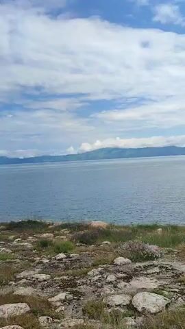 The video captures a serene natural landscape dominated by a vast body of water under a bright, partly cloudy sky. The foreground features a rocky, uneven terrain covered with sparse vegetation, including dry grasses and some purple wildflowers. A simple, open-sided shelter with a translucent roof and a bench inside is visible on the left side of the frame.

As the video progresses, a person wearing dark clothing, possibly a jacket and pants, appears from the right side of the frame and walks towards the water. They are seen near a small tree and some items on the ground. Further in the distance, across the water, a low-lying shoreline is dotted with structures, suggesting the presence of a settlement. The background is filled with rolling hills or mountains that fade into the horizon. The overall atmosphere is tranquil and scenic, with the gentle movement of clouds and the expansive water contributing to the peaceful ambiance.