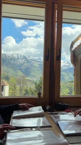 Two individuals are seated at a wooden table inside what appears to be a restaurant or cafe in Epagny, Switzerland. Both are engaged in looking at open menus laid out before them. One person's hands are visible on the left, resting on a menu, while another's hands are on the right, in the process of turning a page.

A large, multi-paneled window with a light brown wooden frame dominates the background, offering a stunning panoramic view. Beyond the window, a majestic mountainous landscape unfolds under a bright blue sky dotted with fluffy white clouds. The distant mountains feature snow-capped peaks, transitioning to lush green, tree-covered slopes and valleys. A small village with houses is visible nestled in the valley below, adding to the picturesque scenery. The weather appears to be clear and sunny.

On the table, a wooden pepper grinder is visible between the menus, and a black camera can be seen near the menu on the right. The audio captures a conversation in French, with audible words like "Chanterelle" and "grillé," suggesting a discussion about food choices from the menu.