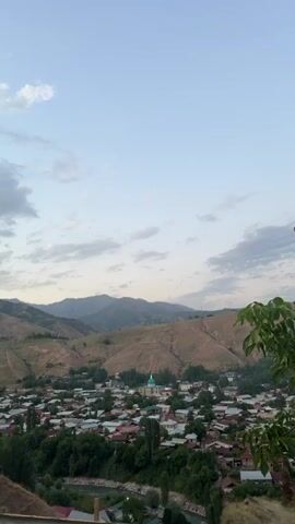 A high-angle, static shot reveals a village nestled in a valley between rolling hills. The sky above is a pale blue, with scattered white clouds. The village features numerous houses with red tile roofs, surrounded by lush green trees. A prominent building with a turquoise dome stands out in the center of the village. The hillsides are covered in dry grass and sparse vegetation, with visible dirt paths winding up their slopes. In the foreground, the leaves of a green tree partially obscure the view. The overall impression is a tranquil, rural landscape.