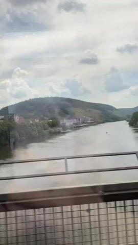 The video is filmed from the perspective of someone on a moving train, looking out the window. A wide river stretches into the distance, bordered by lush green hills on either side. On the left bank, a small town with white buildings and red roofs is visible, nestled amongst trees. The sky above is a mix of blue and white clouds, suggesting a partly cloudy day. A metal railing runs across the bottom of the frame, indicating the presence of a bridge or a viewing platform. The train's movement causes the landscape to appear slightly blurred, adding to the sense of travel.