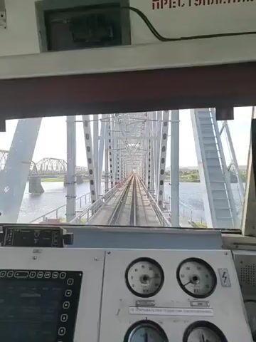 This video shows a first-person view from the cabin of a train as it travels across a large railway bridge. The camera is positioned behind the control panel of the train, looking out through the front window. The bridge is made of white metal trusses and spans a wide river. The tracks run straight across the bridge, disappearing into the distance. On either side of the bridge, there is water, and in the background, the landscape is green with trees. The sky is blue with some scattered clouds, suggesting it is daytime. In the far distance, another bridge can be seen. The control panel in the foreground has various buttons, switches, and gauges, but no specific text is legible. The overall impression is one of movement and a scenic journey over a significant engineering structure.