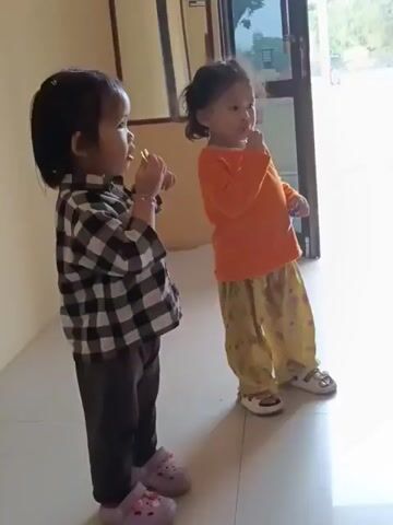 Two young children are standing in a hallway. The child on the left is wearing a black and white checkered shirt, dark pants, and pink Crocs. This child is holding a yellow object in their right hand and appears to be putting it in their mouth. The child on the right is wearing an orange long-sleeved shirt, yellow patterned pants, and white sandals. This child is holding a small object in their right hand and appears to be bringing it to their mouth. The children are standing on a light-colored tiled floor. Behind them is a wall and a doorway with a glass pane. The lighting suggests it is daytime.
