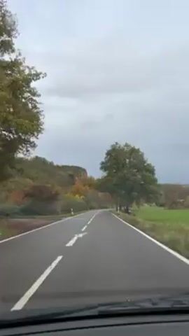 The video shows a car driving down a two-lane road with trees on both sides. The leaves on the trees are changing color, indicating it is autumn. The sky is overcast. The road is wet, suggesting recent rain. There are hills in the background. A white arrow is painted on the road indicating the direction of travel. The video is taken from the perspective of the driver.