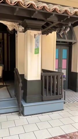 The video shows a panning shot of a seemingly empty shopping mall. The camera starts by focusing on a section with colorful doors and decorative pillars, then pans to the right to show a few people walking through the mall. A young girl and an adult male are seen walking together, followed by a woman with pink-dyed hair. Further down the mall, a man wearing a white baseball cap and a dark hoodie is walking away from the camera. The mall appears to have a tiled floor and some store windows are covered with metal gates. Sunlight streams through the large windows of the mall, casting shadows on the floor.