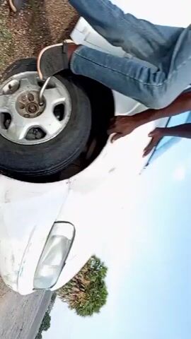A person is shown working on a flat tire of a white vehicle on the side of a road. The vehicle's bumper is visible, and it appears to have some damage. The tire is lying on the ground next to the car, and the person is using a lug wrench to remove the lug nuts. The surroundings consist of a roadside with gravel, grass, and a road surface.