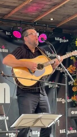 A man wearing glasses and a black short-sleeved shirt is playing an acoustic guitar and singing into a microphone. He is standing on a stage in front of a black backdrop with the words "BAKGården" repeated in a pattern. There are pink lights above him. A music stand is in front of him with sheet music. To the right of the man, a portion of a Yamaha keyboard is visible. The man is holding the guitar with his left hand on the neck and strumming with his right hand. His mouth is open as he sings.